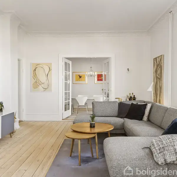 Grå sofa står i lys stue med trægulv, foran fladskærm på lavt skab. Moderne kunst hænger på væggene. Spisebord ses gennem franske døre i baggrunden.