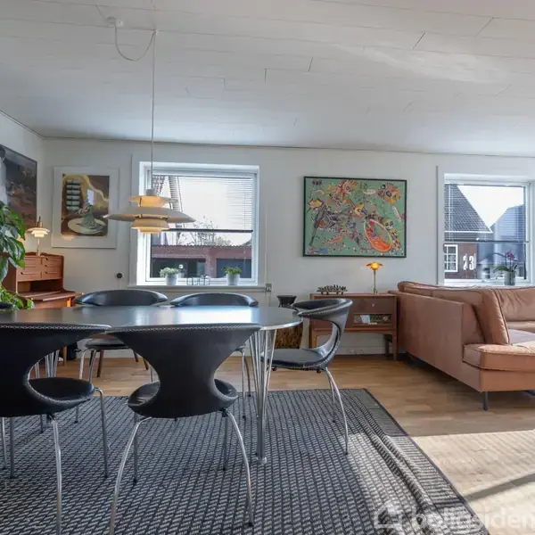 Et moderne opholdsrum med sort spisebord og stole, brun lædersofa, farverige malerier på væggen. Rummet er lyst med store vinduer, og der er en stor tæppe på gulvet.
