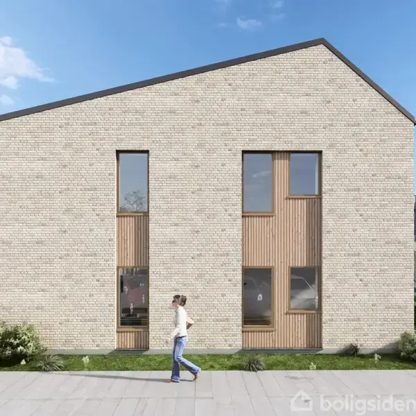 En person går foran en moderne bygning med murstensvægge og høje vinduer. Omgivet af en have og beliggende i et boligområde under en blå himmel.