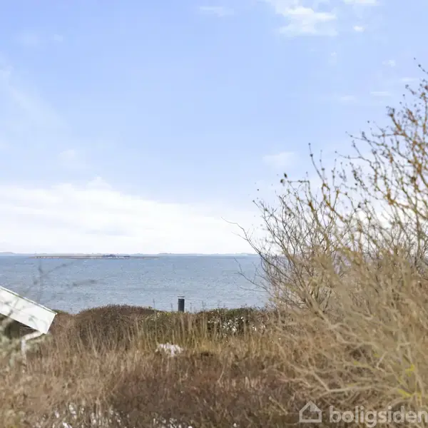 Hus med skråt tag står stille ved en kyst, omgivet af tørre buske, med udsigt over en rolig sø og en blå himmel med skyer.
