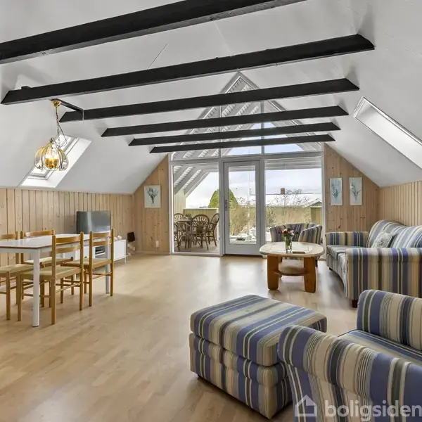 Stue med stribede møbler placeret nær glasdøre, der fører til en balkon. Et spisebord står til venstre, omgivet af trægulve og lyse vægge med træpaneler.