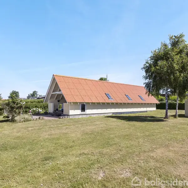 Et rødt tegltag på et langstrakt hus med flere skråvinduer. Huset står på en velplejet græsplæne omgivet af træer og små bygninger i et solrigt område.