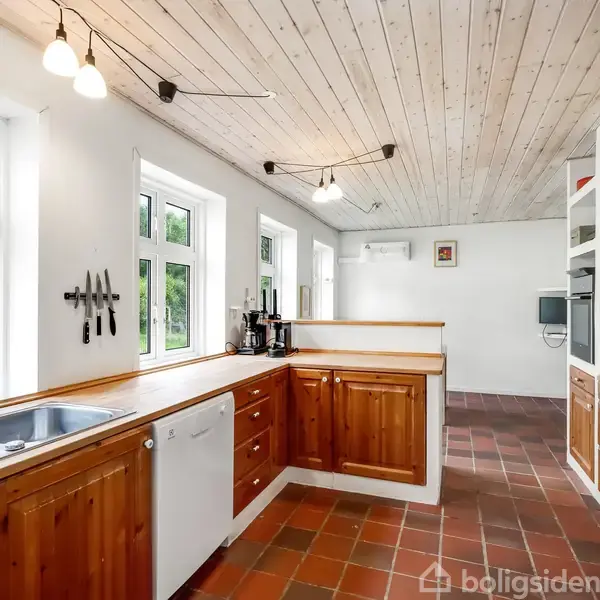 Køkken med træskabsfronter og dobbeltvask i rustfrit stål. Køkkenmaskiner placeret ved vinduerne. Omgivet af lyse vægge og loft med indbygget belysning, klinkegulv i terrakottafarve.