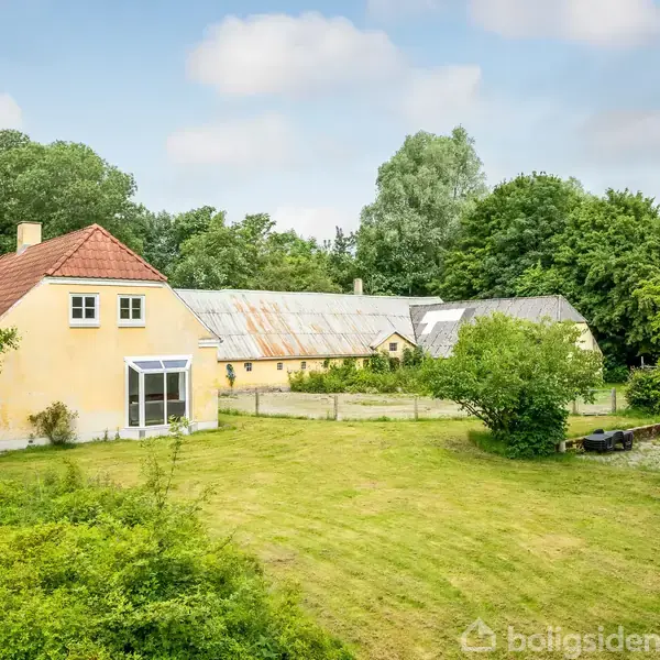 Gul bygning med rødtag står i en have med græsplæne og træer. En lang, grå udhusbygning ses i baggrunden omgivet af frodig vegetation. Skyer ses på himlen.