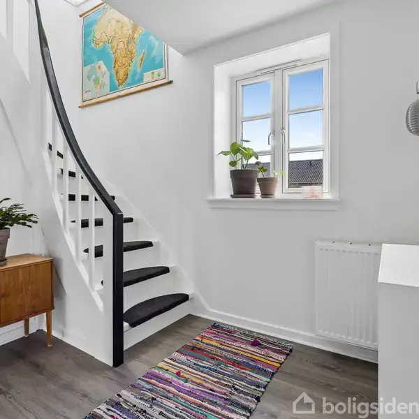 Trappe spiralformet fører fra stueetagen til overetagen. Under et vindue står potteplanter, og et farverigt tæppe pynter det grå gulv. En verdenskortplakat hænger på væggen.
