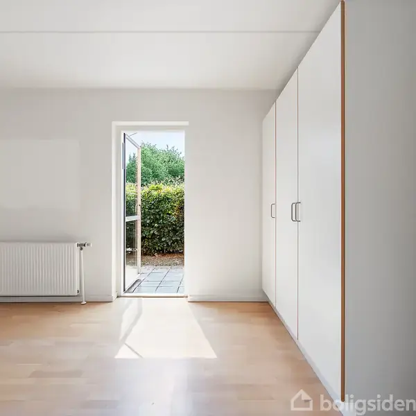 Et lyst værelse med trægulv har et åbent vindue, der fører til en terrasse med udsigt til en grøn hæk. Der er indbyggede skabe til højre og en radiator til venstre.