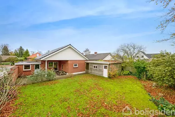 En rød murstensvilla står i en velholdt have, hvor efterårsblade dækker græsset. Huset har en tagterrasse og en lille, skur ved siden af. Roligt nabolag med træer.