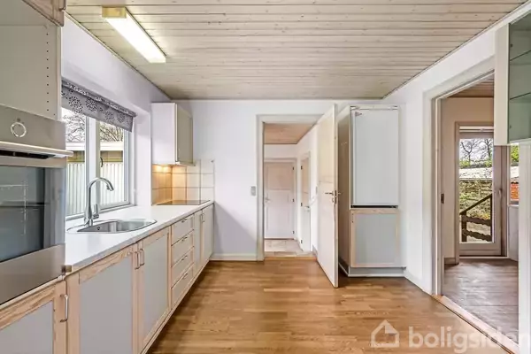 Moderne køkken med en hvid bordplade og stålvask. Der er skabe med trædetaljer og et vindue, der giver udsigt til en gård. Der er adgang til andre rum gennem åbne døre.