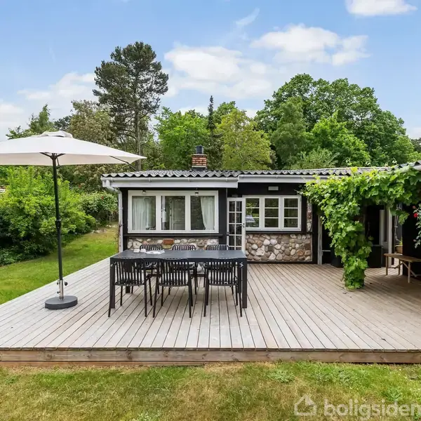 En træterrasse med et bord og stole under en parasol i en frodig have. Huset i baggrunden er delvist dækket af grønne planter.
