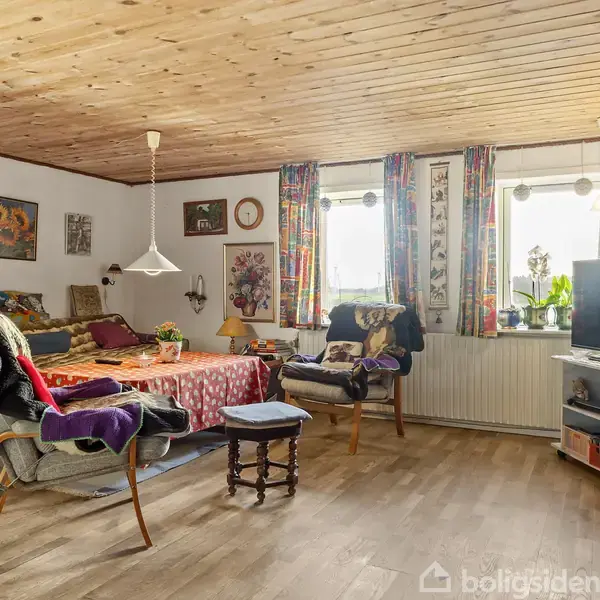 Et velindrettet rum med en seng dækket af et rødt tæppe. To lænestole er placeret ved siden af et lille bord. Væggene er pyntet med kunstværker, og lyse gardiner hænger ved vinduerne. En reol med en TV står ved siden af.