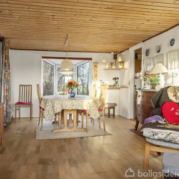 Stue med spisebord dækket med dug og vase med blomster, omgivet af træstole. Hjørnet har en behagelig lænestol med en rød pude, hvorpå der står "I Love You".