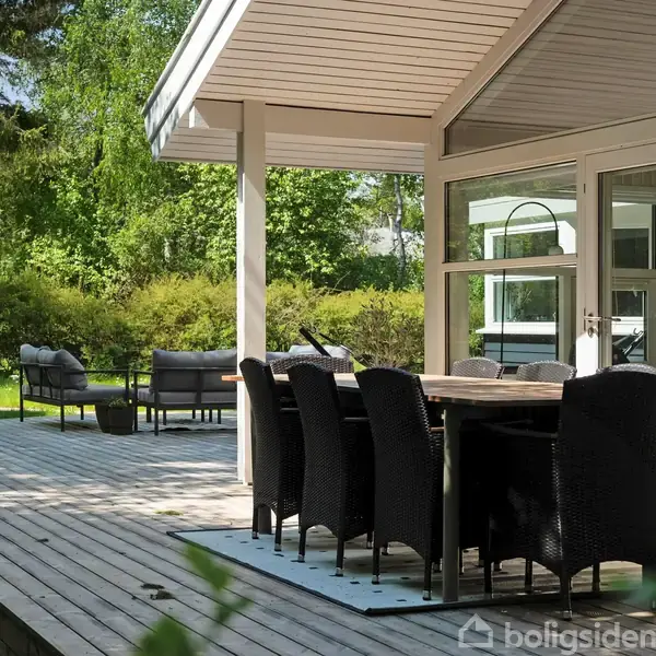 En terrasse med spisebord og stole står under et halvtag. Terrassepladsen er omgivet af frodig grøn beplantning og træer, hvilket skaber en hyggelig udendørs atmosfære.