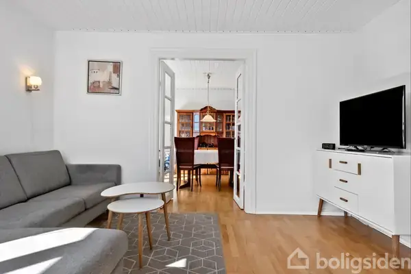 En grå sofa står i stuen ved siden af to små borde. Et tæppe pryder gulvet. Fjernsynet står på en kommode, og spisestuen ses gennem en døråbning.