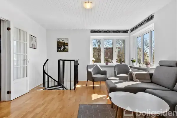 Stue med grå sofa og bord står ved siden af store vinduer med udsigt til træer. En sort spiraltrappe fører nedad. Lyse trægulve og hvidt loft skaber en lys atmosfære.