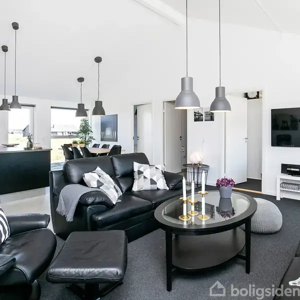 Sort lædermøblement omkring et glasbord med lys, i en moderne stue. Der er fladskærms-TV på væggen, elegante lamper hænger fra loftet, og rummet har lyse, åbne vinduer.