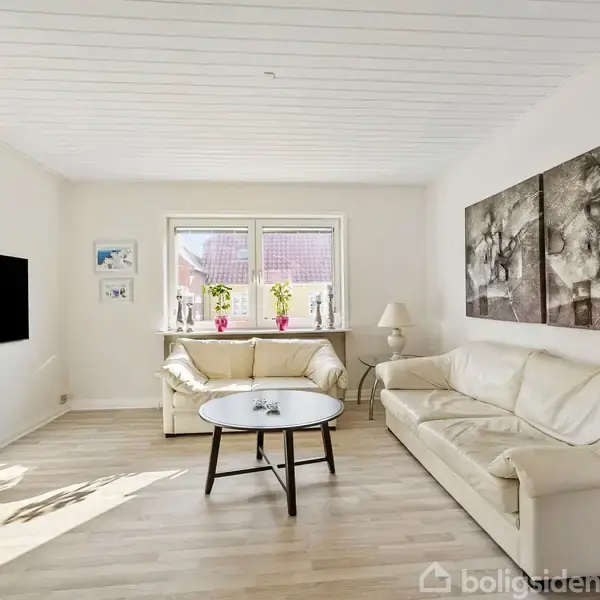 Stue med hvid sofa og lænestol, sofabord foran; vægkunst hænger til højre. TV er monteret til venstre. Store vinduer med udsigt og planter. Lys trælaminatgulv.