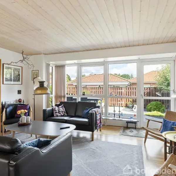 En stue med sorte lædersofaer og stole arrangeret omkring et sofabord. En reol fyldt med bøger står til venstre. Store vinduespartier åbner mod en terrasse og have.