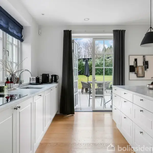 Et moderne køkken med hvide skabe og to sort-hvide lamper over en central ø. Gennem glasdørene ses en terrasse med havemøbler og grøn have i baggrunden.