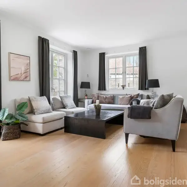 Stue med moderne sofaer arrangeret omkring et lavt sofabord; store vinduer med lange gardiner lyser rummet op; indrettet med planter, lamper og vægkunst, skaber en hyggelig atmosfære.