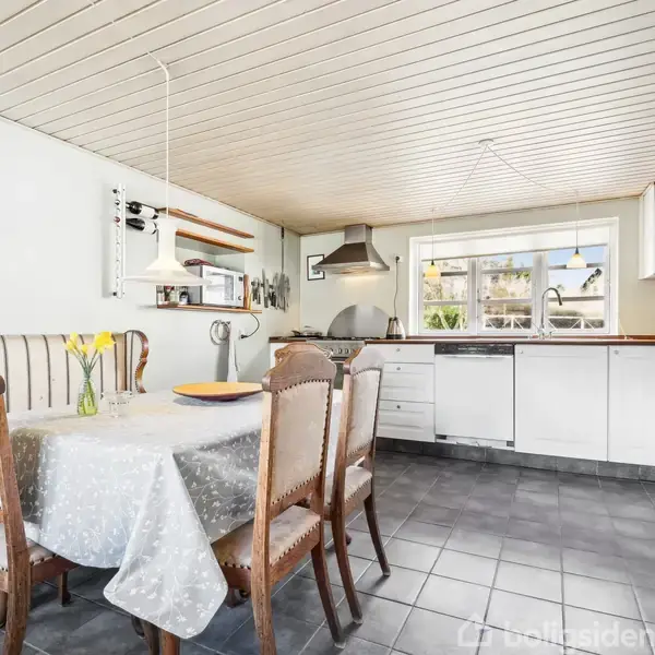 Et rustikt køkken med et træbord og træstole med stofsæder. Bordet har en dug med blomstermønster og en vase med blomster. Køkkenet er lyst med hvide skabe og et vindue.