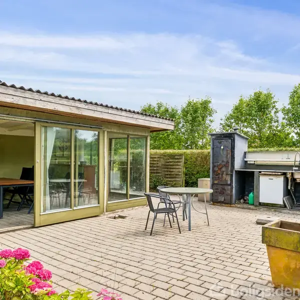 En terrasse med fliser har et bord og stole, en grillplads i baggrunden og en delvist lukket veranda. Omkranset af grønne buske og træer, med blomster i forgrunden.