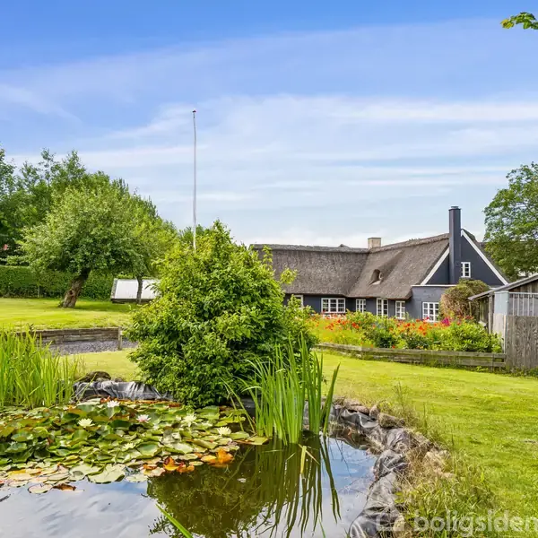 Et stråtækt hus ligger i en frodig have med en dam, omgivet af græsplæne og træer. Et drivhus ses til venstre, indrammet af grønne buske og blomster.