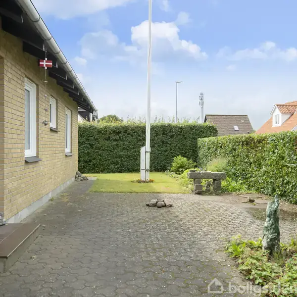 Gulstenshus med hvid dør, der står stille ved en flisebelagt gangsti. En flagstang med et dansk flag står i en velplejet have omgivet af grøn hæk og nabohuse.