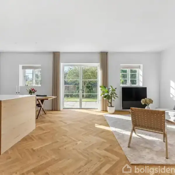 Et moderne, lyst rum med en køkkenø til venstre, en grå sofa og stol til højre, og glasdøre i baggrunden, der fører ud til en have. Trægulv i sildebensmønster.