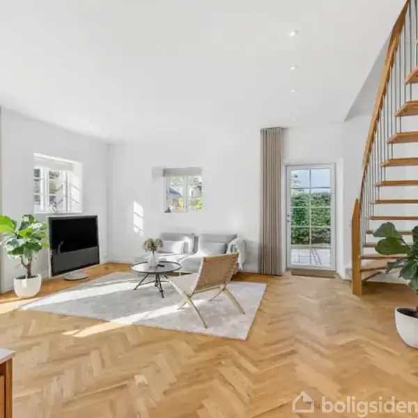 En lys stue med en hjørnesofa og to lænestole centreret omkring et rundt sofabord. En stor fladskærm står til venstre. Trappen til højre leder opad, omgivet af planter.