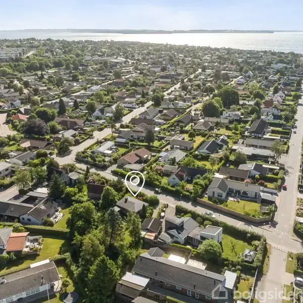 Boligområde set fra oven med et hav af enfamiliehuse og veje. Huset modtager markering med en ikon, der fremhæver det blandt de mange grønne haver og den nærliggende kystlinje.