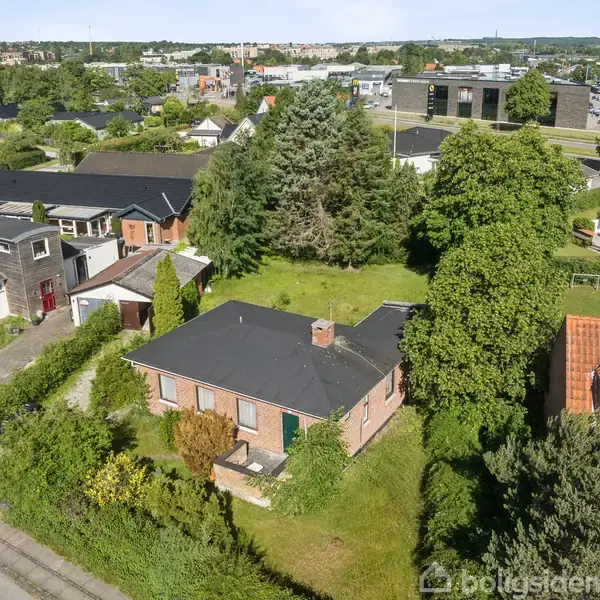 Et enfamiliehus med mørkt tag ligger i en grøn have omgivet af buske og træer. Omkring huset ses flere lignende boliger og veje i et beboelseskvarter.