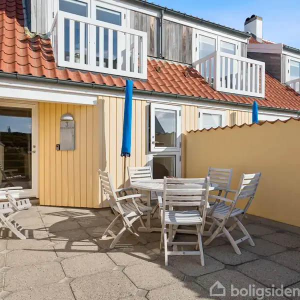 Terrasse med hvide havemøbler er placeret foran et gult hus med rødt tegltag. Blå parasoller står oprejst. Murstensvægge omgiver området. Der er døre til boligen i baggrunden.