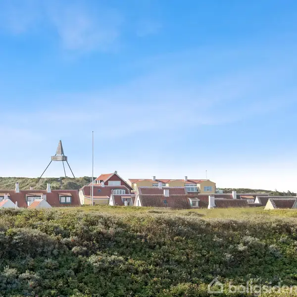 Rækkehuse står stille i en klynge med en høj klokkestruktur i baggrunden og omgivet af en lav grøn høj, under en skyfri blå himmel.