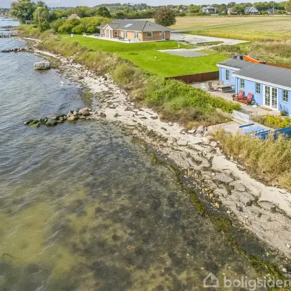 Blåt hus står stille ved en snoet kystlinje ved havet, omgivet af grønne græsarealer og en anden bygning længere væk, under en klar himmel.