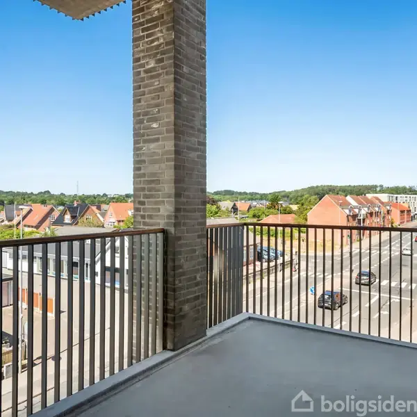 Balkon med metalgelænder tilbyder udsigt over en byvej med kørende biler, omkringliggende huse og grønne træer i baggrunden under en klar blå himmel.