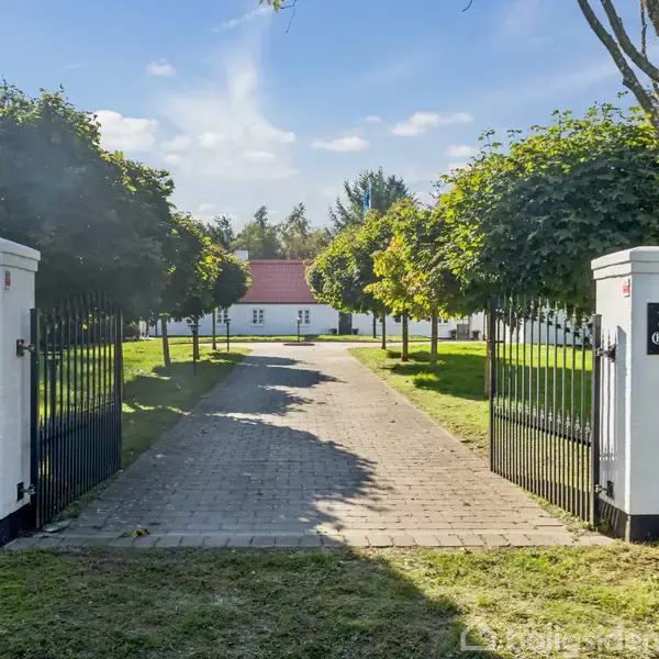 To søjler med skilte "Anno 1900" og "Rita Guardi" flankerer en åben smedejernsport, der fører til en stenbelagt sti omgivet af træer og græs, med et hus i baggrunden.