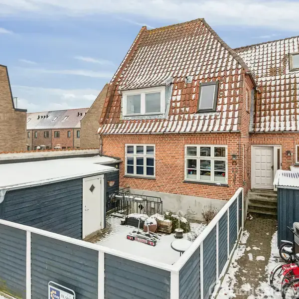 Et rødt murstenshus med skrå tagbeklædninger dækket af sne. En lille gårdhave med cykler og et skur ses i forgrunden. Omkringliggende bygninger danner en bymæssig kontekst.