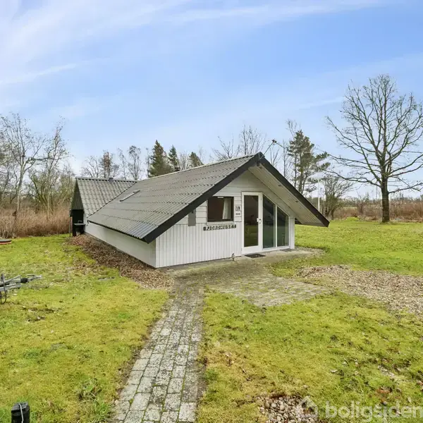 Et hvidt hus med skråtag står på en græsbevokset grund omgivet af træer. En stenbelagt sti fører til hoveddøren. Skilt med teksten "BJØRNSON" over døren.