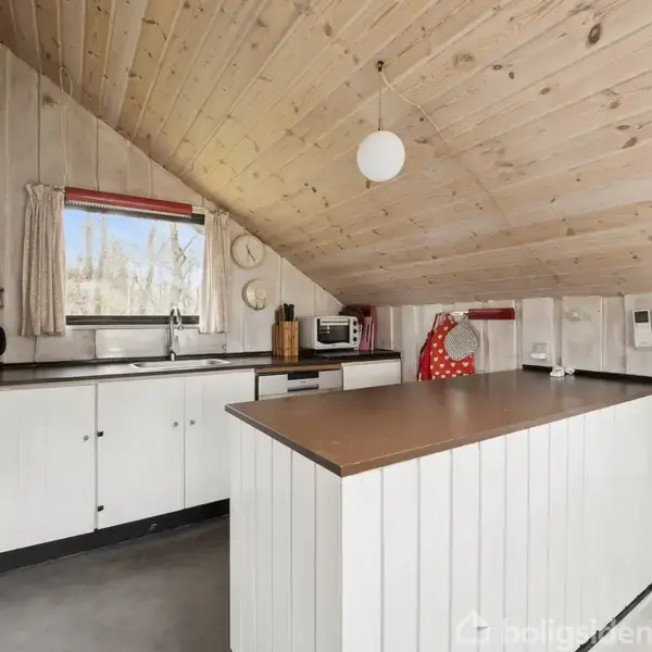Køkken med trævægge og skråt loft, indeholder hvidt skab, komfur, emhætte og køkkenø med brun bordplade. Et vindue giver udsigt, og en krog med rødt viskestykke tilføjer farve.