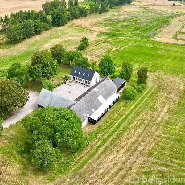 Landbrugsejendom omgivet af åbne marker og grønne træer. Bygningerne danner en gårdsplads, med hovedbygningens mørke tag synligt. Ingen tekst fundet.