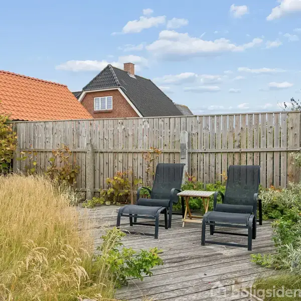 To sorte liggestole står på en træterrasse ved et lille bord. Omkring er der højt græs, blomster og hegn, mens et hus med rødt tag er synligt i baggrunden.