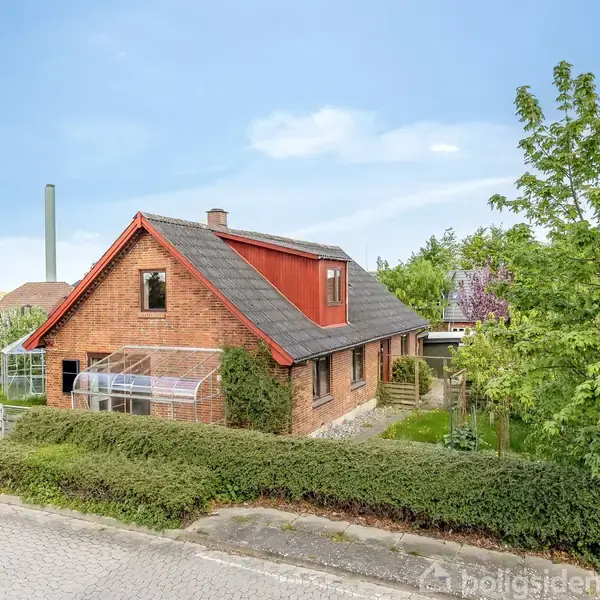 Et rødt murstenshus står stille omgivet af frodige grønne træer og buske i et boligområde. Husets tag er skråt, med en lille drivhussektion på siden.