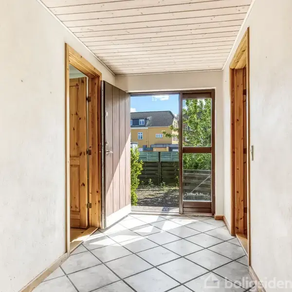 En lys gang med hvide fliser på gulvet leder til en glasdør. Faciliteterne mødes med trædøre, og udenfor vises en have med et hus i baggrunden.