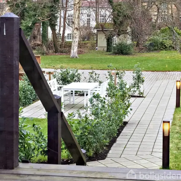 En flisebelagt sti fører gennem en have med lygtepæle langs kanten. En hvid bænk og bord står omgivet af buske. I baggrunden ses træer og en græsplæne.