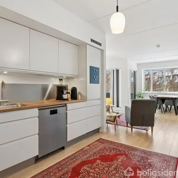 Et moderne køkken med hvide skabe og træbordplade har en opvaskemaskine. En stue er synlig i baggrunden med sofa, stol og spisebord ved store vinduer med udsigt til træer.