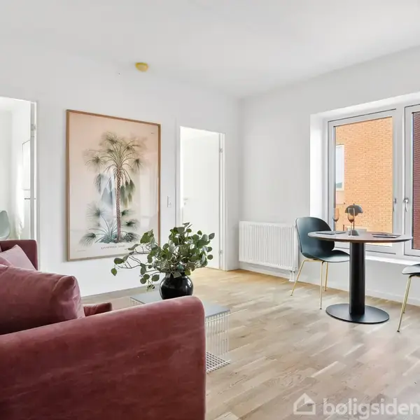 Rød sofa står i et lyst rum med et træplankegulv, hvor der også er et lille bord og to stole ved vinduet. En plante og et stort billede pryder væggen.