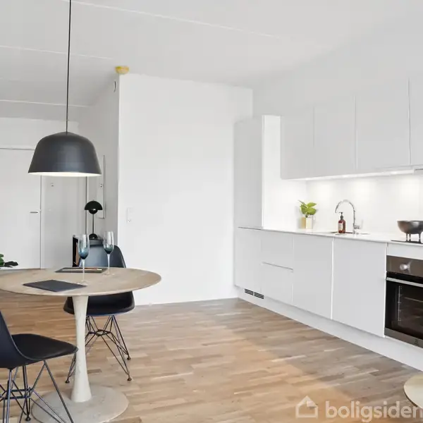 Et moderne køkken-alrum med hvidt køkkenmodul og indbygningsovn. En rund spisebord med to stole står foran, oplyst af en sort pendellampe. Trægulv og vægdekorationer skaber en hyggelig atmosfære.