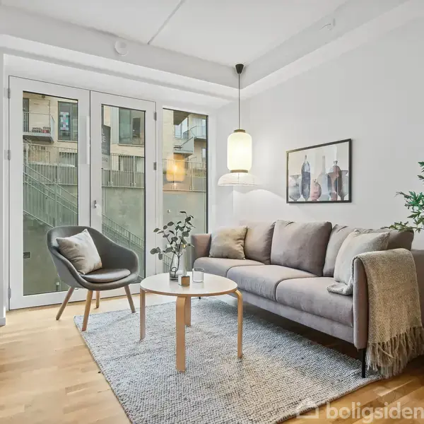 En stue med en grå sofa, et rundt sofabord og en stol. En plante står ved siden af sofaen. Store vinduer viser en bygning udenfor. Et tæppe dækker gulvet.