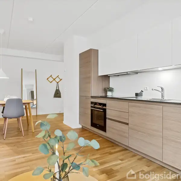 Et moderne køkken med træskabe og sort bordplade står til højre, mens et spisebord med grå stole er placeret til venstre i en lys spisestue.