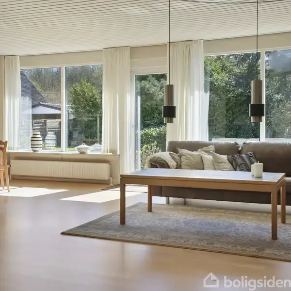 Brun sofa står i solrig stue med stort vindue, lys trægulv og tæppe. Langbord foran sofa, spisebord til venstre, grøn udsigt udenfor.